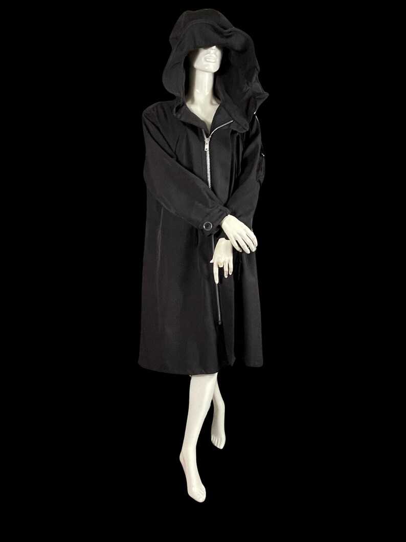 May include: A long black hooded coat with a silver zipper closure. The coat has long sleeves and a button detail on the cuffs. The mannequin is wearing white gloves and is set against a black background.