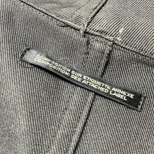 May include: Close-up of gray denim fabric with visible stitching. A black label is attached, displaying the text "COMPOSITION SUR ETIQUETTE ANNEXE COMPOSITION: SEE ATTACHED LABEL".