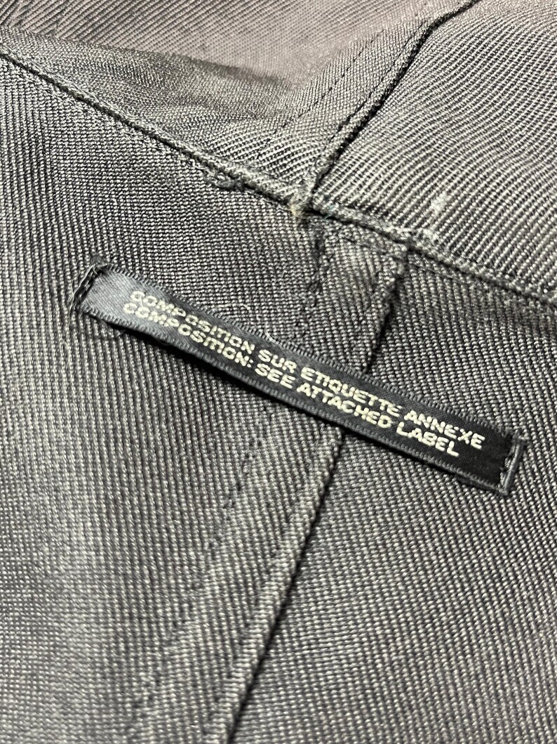 May include: Close-up of gray denim fabric with visible stitching. A black label is attached, displaying the text "COMPOSITION SUR ETIQUETTE ANNEXE COMPOSITION: SEE ATTACHED LABEL".