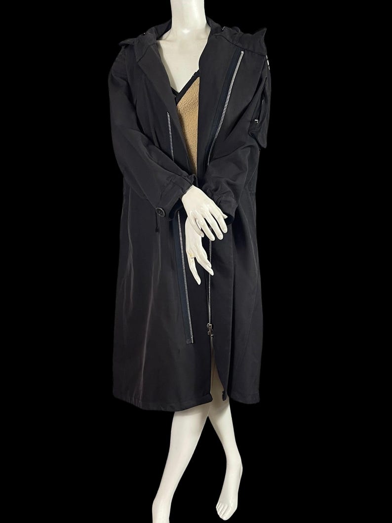 May include: A long black coat with a hood and silver zippers. The coat has a tailored fit and a sleek, modern design. The coat is shown on a mannequin.