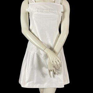 May include: A white, sleeveless dress with a square neckline and wide shoulder straps. The dress has a fitted bodice and a flared skirt, and is displayed on a mannequin against a black background. The dress appears to be made of a smooth, possibly satin-like fabric.