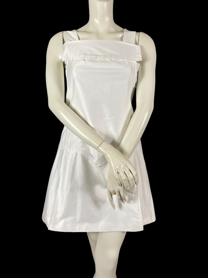 May include: A white, sleeveless dress with a square neckline and wide shoulder straps. The dress has a fitted bodice and a flared skirt, and is displayed on a mannequin against a black background. The dress appears to be made of a smooth, possibly satin-like fabric.