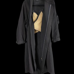 May include: A long black coat with a hood and a zipper closure. The coat has a gold-colored lining visible in the open front. The coat is hanging on a black hanger against a black background.