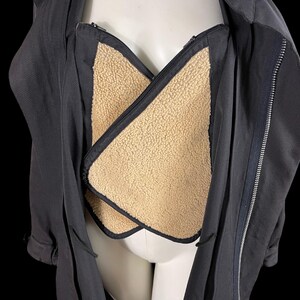 May include: A black jacket with a beige, fuzzy lining. The jacket has a zipper closure and a collar. The lining is visible in the chest area. The jacket is displayed on a mannequin against a black background.