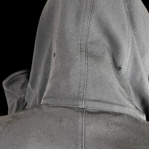 May include: Close-up of a dark gray hooded jacket. The hood is up, revealing the back of the jacket and the hood's construction. The fabric has a subtle diagonal weave, and the hood has two small eyelets.