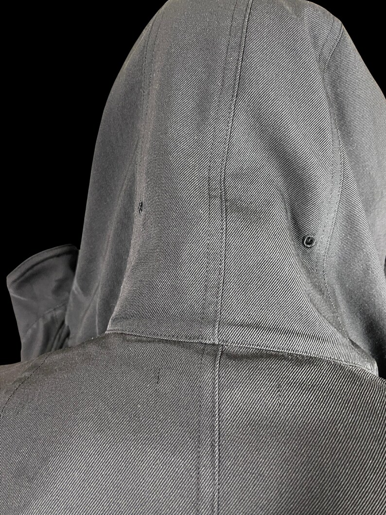 May include: Close-up of a dark gray hooded jacket. The hood is up, revealing the back of the jacket and the hood's construction. The fabric has a subtle diagonal weave, and the hood has two small eyelets.
