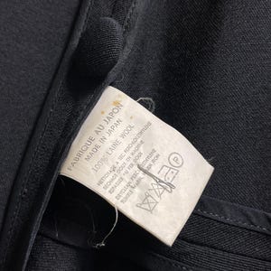 May include: Close-up of a black garment with a tag attached. The tag reads "FABRIQUE AU JAPON MADE IN JAPAN 100% LAINE WOOL" with care instructions. The garment's fabric appears to be wool.