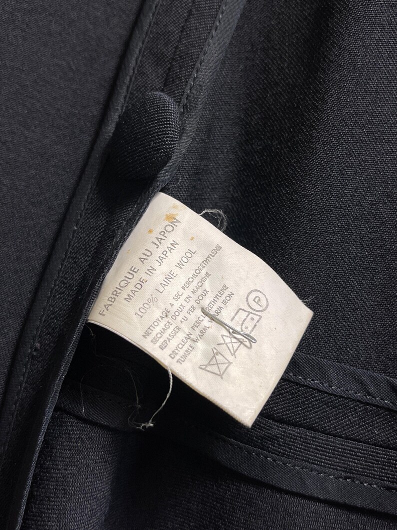 May include: Close-up of a black garment with a tag attached. The tag reads "FABRIQUE AU JAPON MADE IN JAPAN 100% LAINE WOOL" with care instructions. The garment's fabric appears to be wool.
