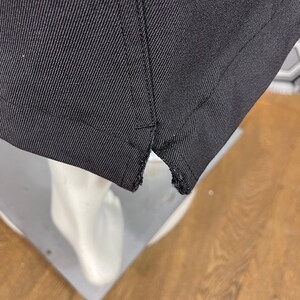 May include: Close-up of a black fabric, possibly a garment, showing the hem and stitching details. The fabric appears to be a sturdy material, with a visible weave. The hem is slightly frayed.