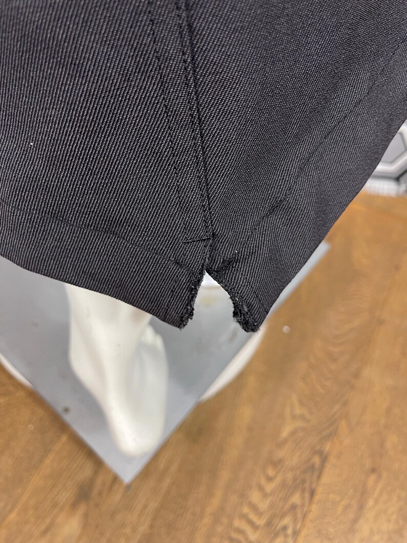 May include: Close-up of a black fabric, possibly a garment, showing the hem and stitching details. The fabric appears to be a sturdy material, with a visible weave. The hem is slightly frayed.