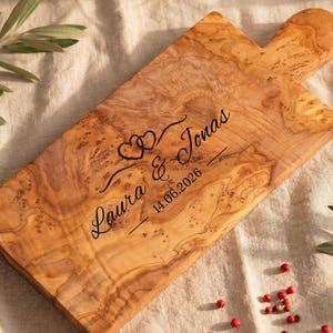 May include: Wooden cutting board with a handle, engraved with the names "Laura & Jonas", two hearts, and the date "14.06.2026". The board has a natural wood grain pattern and is surrounded by olive branches and red peppercorns.