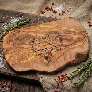 May include: A wooden cutting board with a natural wood grain pattern. The board has an engraved floral design and text that reads "HERZLICH WILLKOMMEN in unserem Zuhause Familie M&uuml;ller Seit 2021". Rosemary sprigs and spices surround the board.