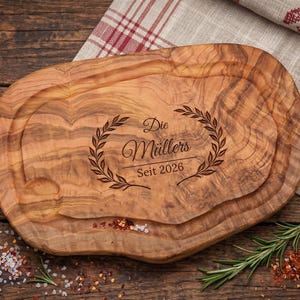 May include: A wooden cutting board with a natural wood grain pattern. The board has a carved-out section and features the text "Die Müllers Seit 2026" within a laurel wreath design. Rosemary sprigs and spices are scattered around the board.