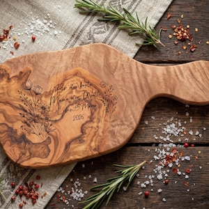 May include: Wooden cutting board with a handle, featuring engraved floral designs and the text "F&uuml;r die beste Mama der Welt Lukas & Mia 2026". The board is light brown with a natural wood grain pattern. Rosemary sprigs and spices surround the board.