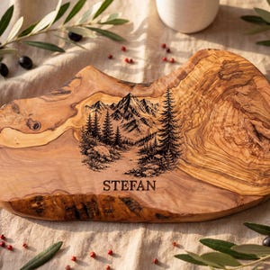 Puede incluir: Una tabla de servir de madera de olivo con bordes irregulares y naturales, que presenta un grabado láser negro de una cordillera con pinos y un sendero forestal. El nombre STEFAN está grabado en una fuente negrita sin remates bajo la escena.