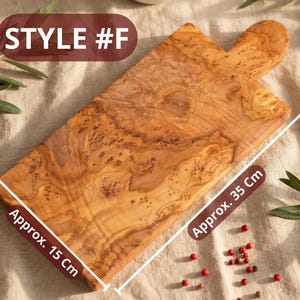 May include: Wooden cutting board, style #F, with a handle. The board is approximately 15 cm wide and 35 cm long. The wood grain pattern is visible. The background includes olive branches and red peppercorns.