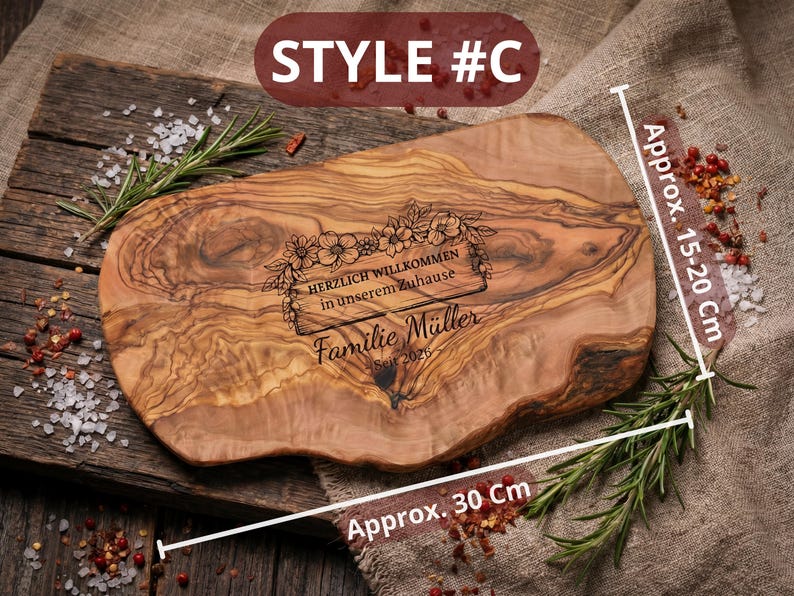 May include: Wooden serving board, approximately 12 inches long and 6-8 inches wide, with engraved text and floral design. The text reads "HERZLICH WILLKOMMEN in unserem Zuhause Familie M&uuml;ller Seit 2026". The board is on a wooden surface with rosemary and spices.