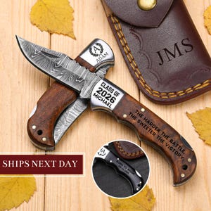 May include: A personalised folding knife with a wooden handle and a Damascus steel blade. The knife is engraved with "CLASS OF 2026 MICHAEL" and "THE HARDER THE BATTLE, THE SWEETER THE VICTORY." A leather sheath with initials is also included.