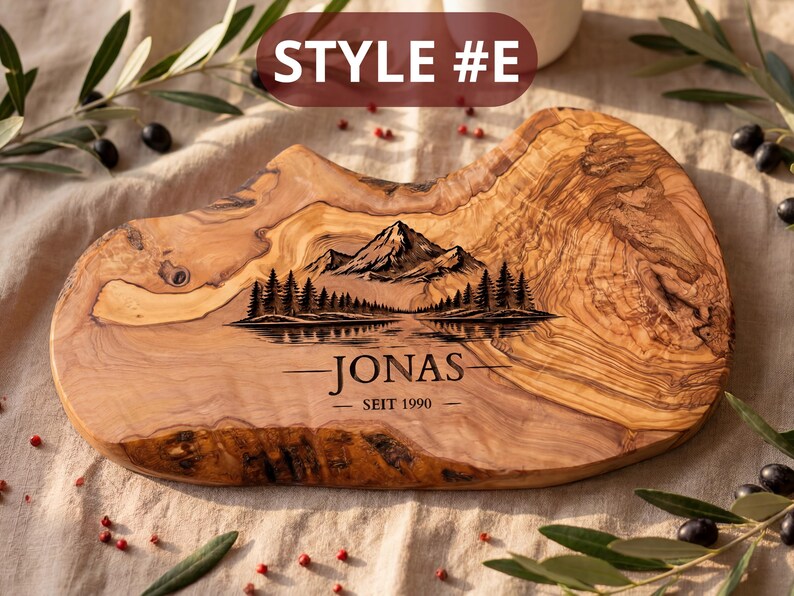 May include: A wooden cutting board with a natural edge, featuring a mountain and lake scene engraving. The board has the text "JONAS SEIT 1990" and "STYLE #E" on it. The board is made of olive wood.