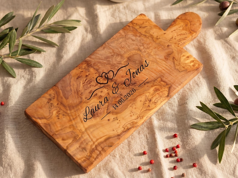 May include: Wooden cutting board with a handle, engraved with the names "Laura & Jonas", two hearts, and the date "14.06.2026". The board has a natural wood grain pattern and is surrounded by olive branches and red peppercorns.