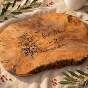 May include: Oval wooden serving board with a natural wood grain. The board features a floral bouquet and butterfly design, along with the text "Beste Mama der Welt" and the names "Lukas & Marie &bull; 2026".