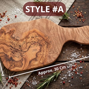 May include: A wooden cutting board with a handle, measuring approximately 12 inches long and 6 inches wide. The board features engraved floral designs and text in German. The text reads "F&uuml;r die beste Mama der Welt" and "Lukas & Mia 2026".