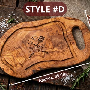 May include: A wooden cutting board with a handle, engraved with "THOMAS KITCHEN" and kitchen utensils. The board is approximately 14 inches long and 8-12 inches wide. The board has a groove around the edge and is made of a light brown wood.