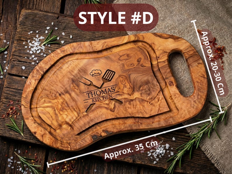 May include: A wooden cutting board with a handle, engraved with "THOMAS KITCHEN" and kitchen utensils. The board is approximately 14 inches long and 8-12 inches wide. The board has a groove around the edge and is made of a light brown wood.