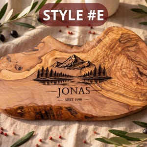May include: A wooden cutting board with a natural edge, featuring a mountain and lake scene engraving. The board has the text "JONAS SEIT 1990" and "STYLE #E" on it. The board is made of olive wood.