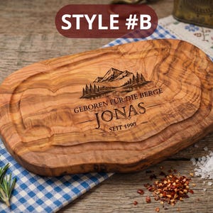 May include: A wooden cutting board with a mountain and forest design, the name "Jonas", and the text "GEBOREN F&Uuml;R DIE BERGE" and "SEIT 1990" engraved on it. The board is oval-shaped with a juice groove. "STYLE #B" is printed at the top.