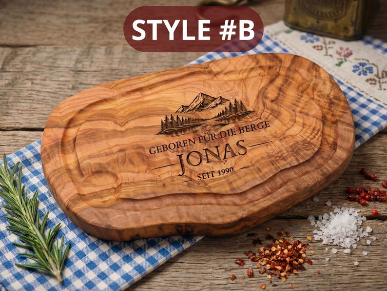 May include: A wooden cutting board with a mountain and forest design, the name "Jonas", and the text "GEBOREN F&Uuml;R DIE BERGE" and "SEIT 1990" engraved on it. The board is oval-shaped with a juice groove. "STYLE #B" is printed at the top.