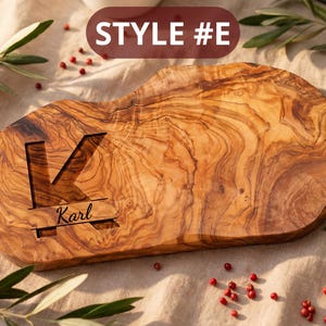 May include: A wooden cutting board with a natural wood grain pattern. The board features a large letter "K" carved into the surface, with the name "Karl" below. The text "STYLE #E" is displayed at the top. The board is surrounded by greenery and red berries.
