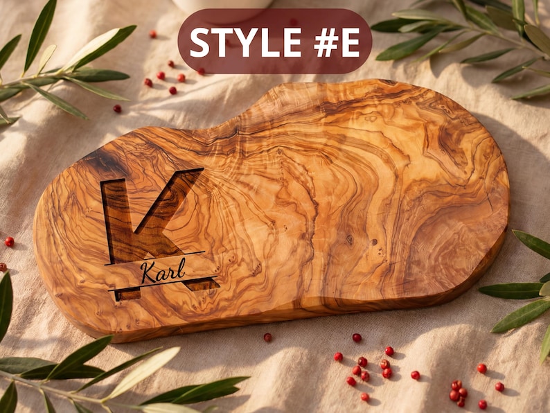 May include: A wooden cutting board with a natural wood grain pattern. The board features a large letter "K" carved into the surface, with the name "Karl" below. The text "STYLE #E" is displayed at the top. The board is surrounded by greenery and red berries.