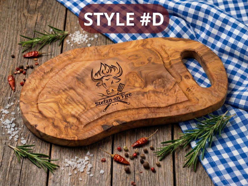 May include: Oval-shaped wooden cutting board with a handle, featuring a carved design of a bull's head and the text "Stefan on Fire." The board is surrounded by rosemary sprigs, red peppers, and salt.