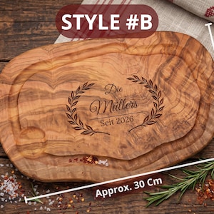 May include: Oval wooden cutting board, approximately 30 cm long and 15-20 cm wide, with a carved groove. The board features engraved text and a laurel wreath design. The text reads "Die M&uuml;llers Seit 2026".
