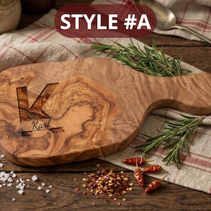 May include: Wooden cutting board with a handle, featuring a carved initial "K" and the name "Karl". The board is made of olive wood and has a natural wood grain pattern. The text "STYLE #A" is at the top. Rosemary sprigs and red pepper flakes are scattered around the board.