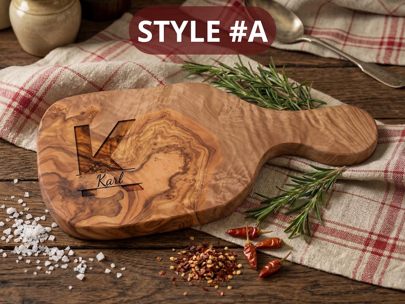 May include: Wooden cutting board with a handle, featuring a carved initial "K" and the name "Karl". The board is made of olive wood and has a natural wood grain pattern. The text "STYLE #A" is at the top. Rosemary sprigs and red pepper flakes are scattered around the board.