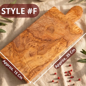 May include: Wooden cutting board, style #F, with a handle. The board is approximately 15 cm wide and 35 cm long. The wood grain is visible. The background includes olive branches and red peppercorns.