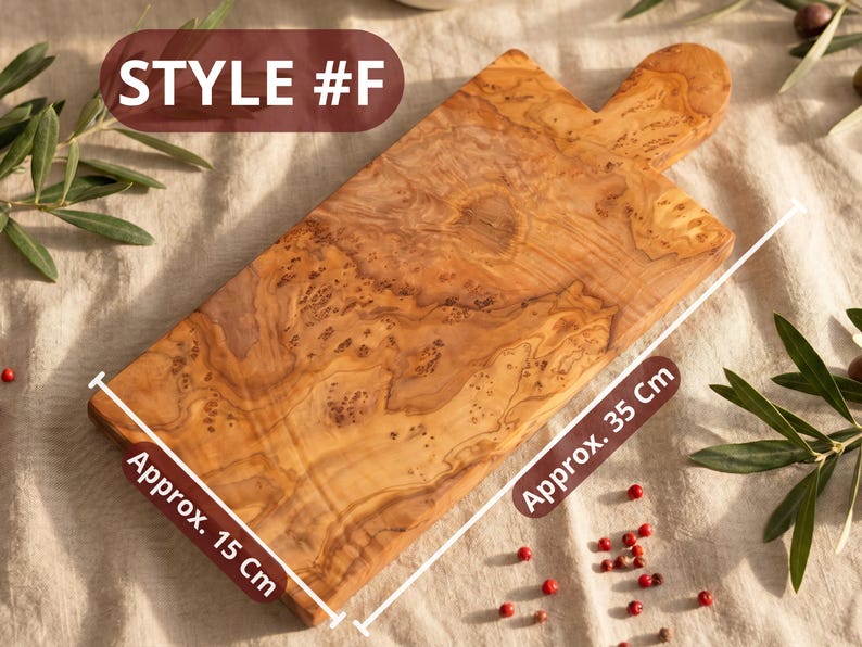 May include: Wooden cutting board, style #F, with a handle. The board is approximately 15 cm wide and 35 cm long. The wood grain is visible. The background includes olive branches and red peppercorns.