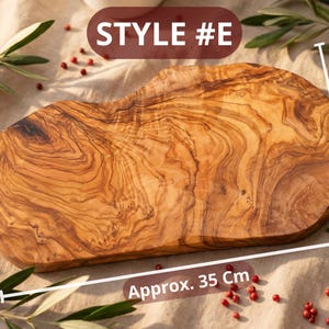 May include: A wooden cutting board with a natural wood grain pattern. The board is approximately 14 inches long and 8-12 inches wide. The text "STYLE #E" is displayed on the top. The board is surrounded by olive branches and red peppercorns.