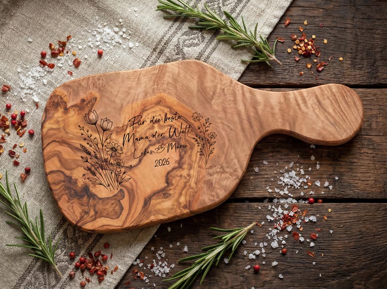 May include: Wooden cutting board with a handle, featuring engraved floral designs and the text "F&uuml;r die beste Mama der Welt Lukas & Mia 2026". The board is light brown with a natural wood grain pattern. Rosemary sprigs and spices surround the board.