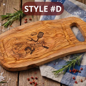 May include: Wooden cutting board with a handle, featuring a carved design with the text "Thomas Kitchen" and kitchen utensils. The board is made of olive wood and has a natural wood grain pattern. The text "STYLE #D" is in the top left corner.