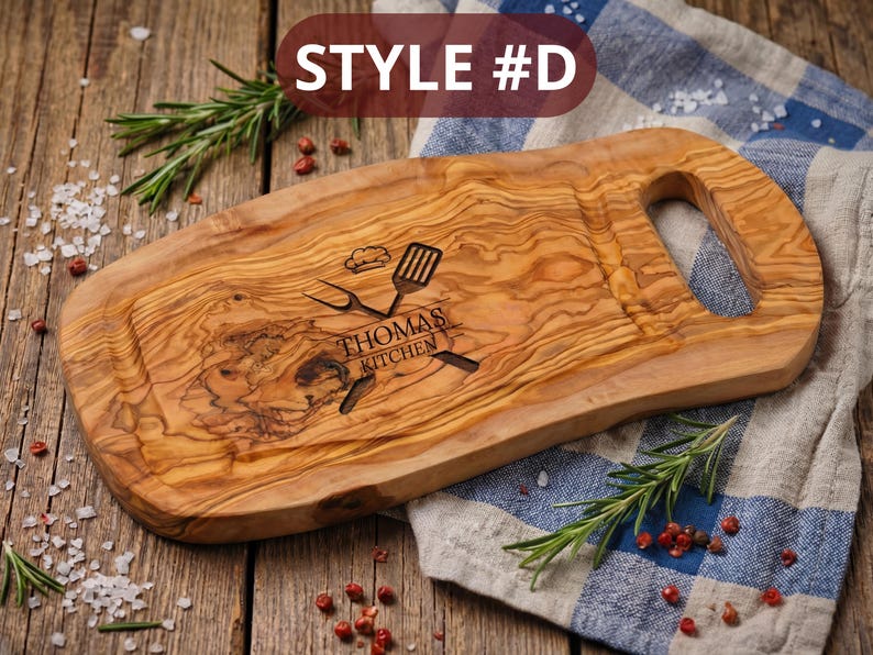 May include: Wooden cutting board with a handle, featuring a carved design with the text "Thomas Kitchen" and kitchen utensils. The board is made of olive wood and has a natural wood grain pattern. The text "STYLE #D" is in the top left corner.