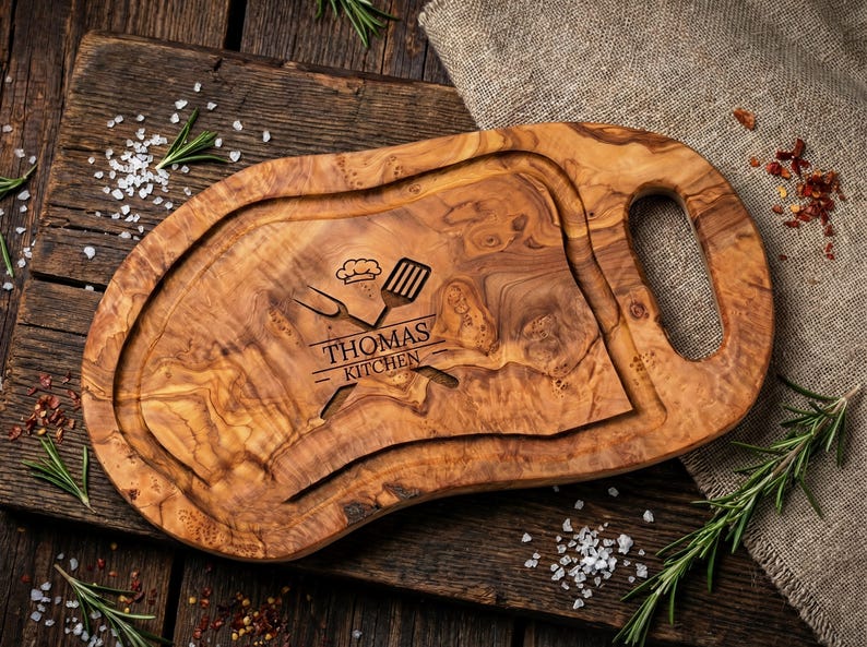 May include: Oval-shaped wooden cutting board with a handle, featuring a carved groove and the text "THOMAS KITCHEN" with chef's tools. The board is made of light brown wood with a natural grain pattern. Rosemary sprigs and salt are scattered around the board.