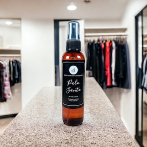 May include: A brown glass bottle with a black label that says "Sweet Comfort Candles LLC Palo Santo Oil & Room Spray" on a light brown countertop. The bottle is in focus, and the background is blurred.