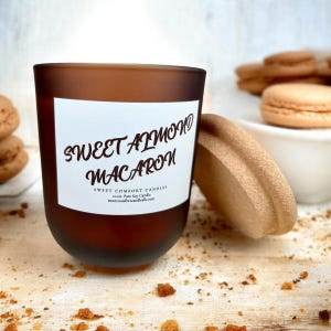 May include: A brown glass candle with the text "Sweet Almond Macaron" on a white label. The candle is next to a tan lid and a small bowl of macarons. The candle is 10 oz. Pure Soy Candle.