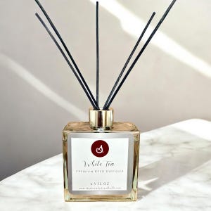 Peut inclure: Un diffuseur de roseaux en verre transparent avec un couvercle doré et des roseaux noirs, rempli d'un diffuseur de roseaux parfumé au thé blanc. L'étiquette indique "White Tea Premium Reed Diffuser 6.5 FL OZ www.sweetcomfortandscents.com".