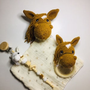 May include: A set of two crocheted brown horse head toys with white markings. One horse head is attached to a wooden teething ring and the other is attached to a wooden rattle with a white crocheted bunny. The toys are on a white fabric with a floral pattern.