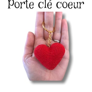 Puede incluir: Llavero de coraz&oacute;n rojo tejido a crochet con una cadena y un anillo dorados. El llavero se sostiene en una mano.