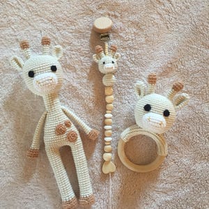 Può includere: Un set di tre giocattoli per bambini all'uncinetto: un peluche a forma di giraffa, un fermaciuccio e un anello per la dentizione in legno. Il peluche giraffa è color crema con macchie e dettagli marroni. Il fermaciuccio ha perline di legno e una testa di giraffa.